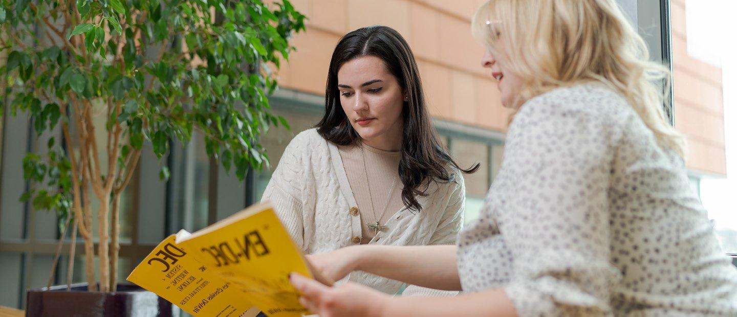 一个女人拿着一本黄色的书，指着一页的一部分给坐在她旁边的另一个女人看.