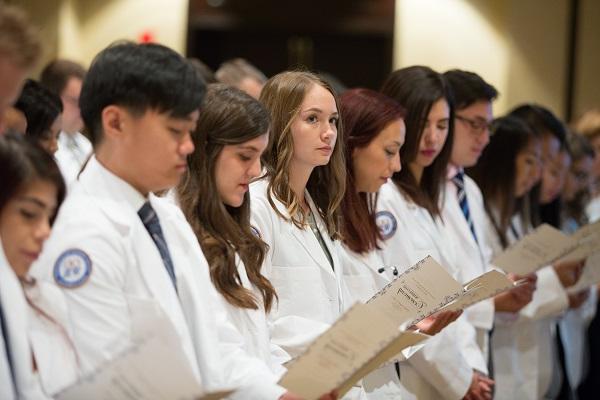 Students stand, holding programs, at the white coat ceremony.