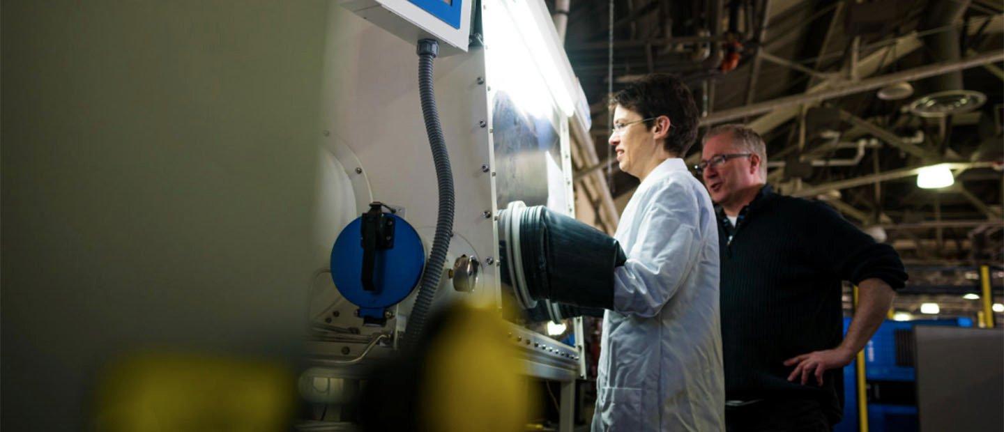 一个穿着白色实验室大褂的女人在一台大型机器前工作，而一个男人在她身后监视.