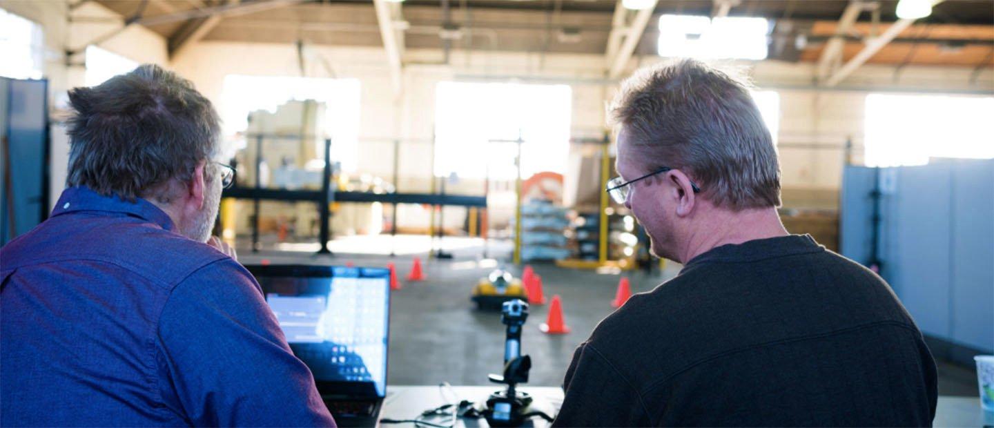 Two men operating a small remote controlled vehicle in a warehouse.