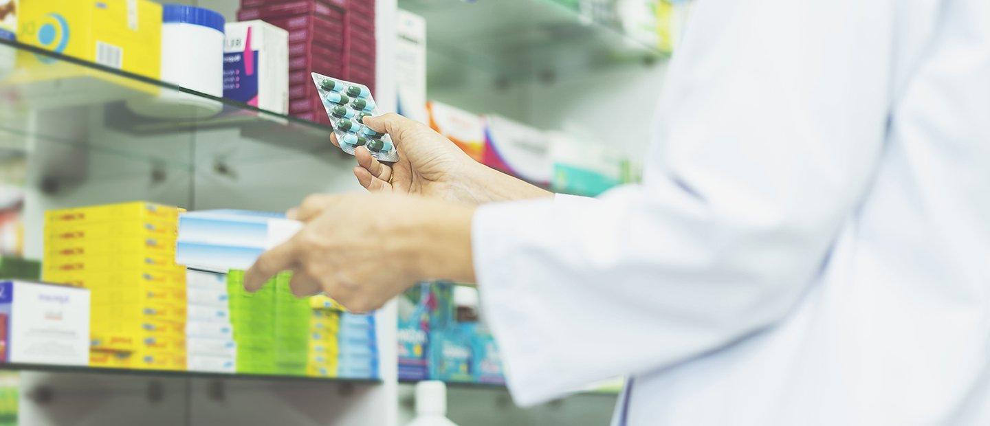 Person in a white lab coat holding a pack of pills in front of a shelf of medicine.