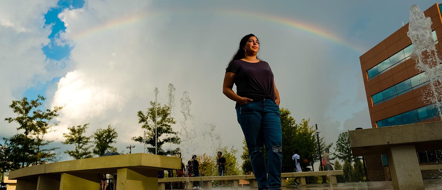 一位年轻女子站在十大菠菜台子的校园里，双手插在口袋里, 身后有彩虹和喷泉.